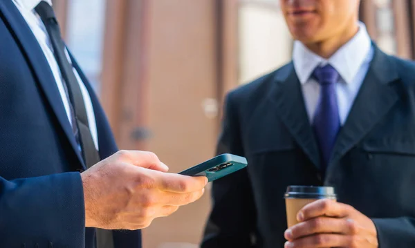 Un homme d'affaires confiant et son collègue utilisent un smartphone devant un immeuble de bureaux moderne. Les investisseurs financiers parlent en plein air. Banques et entreprises. — Photo