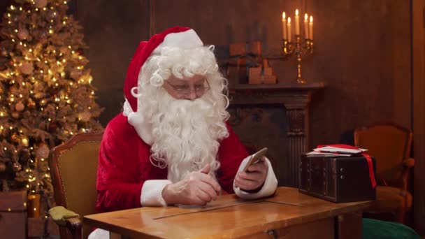 Miejsce pracy Świętego Mikołaja. Wesoły Mikołaj pracuje za pomocą smartfona, siedząc przy stole. Kominek i choinka w tle. Koncepcja świąteczna. — Wideo stockowe