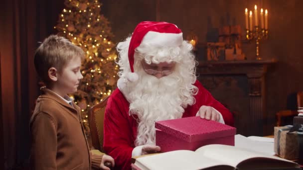 Santa Claus y el niño pequeño. Alegre Santa está trabajando mientras está sentado a la mesa. Chimenea y árbol de Navidad en el fondo. Concepto de Navidad. — Vídeos de Stock