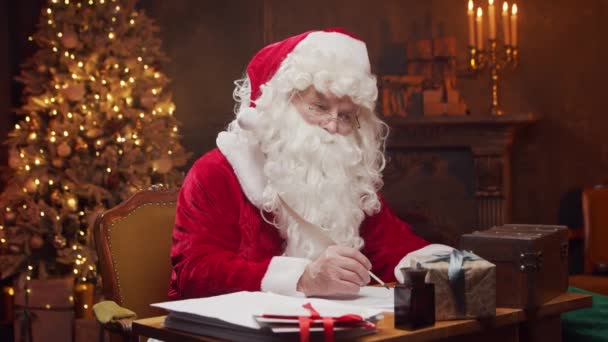 Lugar de trabajo de Santa Claus. Alegre Santa está escribiendo la carta mientras está sentado a la mesa. Chimenea y árbol de Navidad en el fondo. Concepto de Navidad. — Vídeos de Stock
