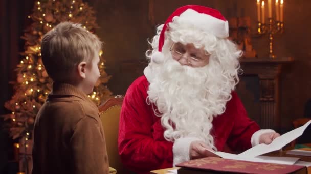 클로스와 쾌활하게 산타는 식탁에 앉아서 일하고 배경은 크리스마스 트리와 벽난로이다 — 비디오