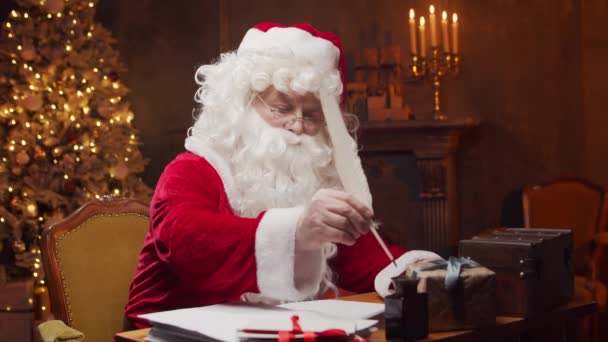 Lugar de trabajo de Santa Claus. Alegre Santa está escribiendo la carta mientras está sentado a la mesa. Chimenea y árbol de Navidad en el fondo. Concepto de Navidad. — Vídeos de Stock