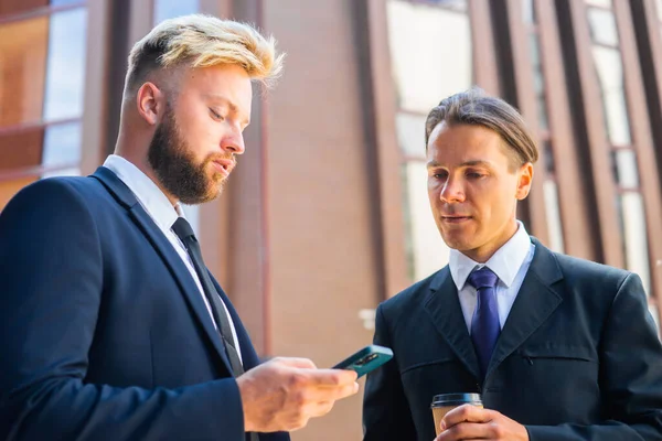 Un homme d'affaires confiant et son collègue utilisent un smartphone devant un immeuble de bureaux moderne. Les investisseurs financiers parlent en plein air. Banques et entreprises. — Photo
