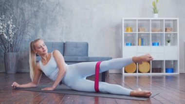 Spor giyim sektöründe genç ve sportif bir kız iç mekanda egzersiz yaparken direniş bandı kullanıyor. Formda ve narin sarışın kadın spor ve spor için geliyor. Sağlık sigortası, yağ yakma ve sağlık..