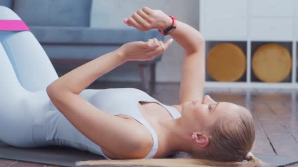 Jong en sportief meisje in sportkleding is met behulp van slimme horloge, terwijl het doen van oefeningen in huis interieur. Fit en slanke blonde vrouw gaat in voor sport en fitness. Gezondheidszorg, vetverbranding en welzijn. — Stockvideo