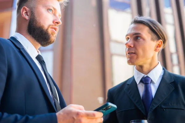 Un hombre de negocios confiado y su colega están usando un teléfono inteligente frente al moderno edificio de oficinas. Los inversores financieros están hablando al aire libre. Banca y negocios. — Foto de Stock