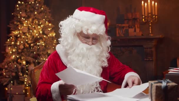 Lugar de trabajo de Santa Claus. Alegre Santa es la lectura de cartas de los niños mientras se sienta a la mesa. Chimenea y árbol de Navidad en el fondo. Concepto de Navidad. — Vídeos de Stock