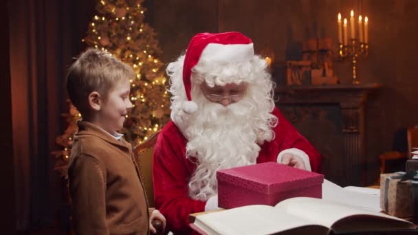 サンタクロースと男の子 陽気サンタはテーブルに座っている間に働いています 背景にある暖炉とクリスマスツリー 伝統的なクリスマスのコンセプト — ストック動画