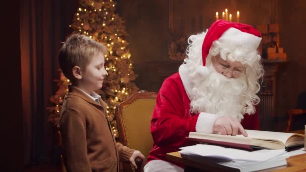 Babbo Natale e il bambino. Allegro Babbo Natale sta lavorando mentre è seduto a tavola. Camino e Albero di Natale sullo sfondo. Concetto Natale. — Video Stock