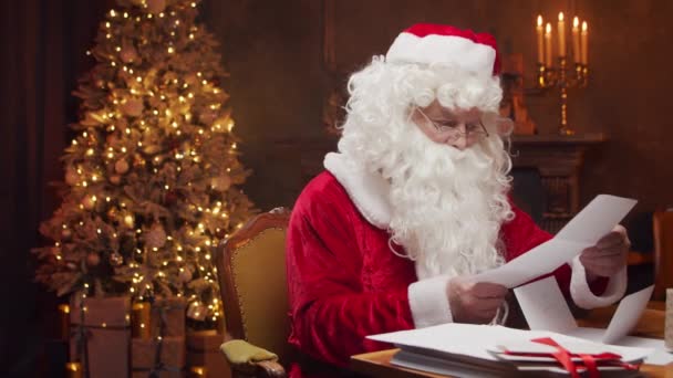 Lugar de trabajo de Santa Claus. Alegre Santa es la lectura de cartas de los niños mientras se sienta a la mesa. Chimenea y árbol de Navidad en el fondo. Concepto de Navidad. — Vídeos de Stock