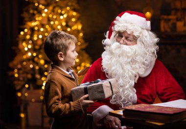 Noel Baba ve küçük çocuk. Neşeli Noel Baba masada otururken çalışıyor. Arka planda şömine ve Noel ağacı var. Noel konsepti.