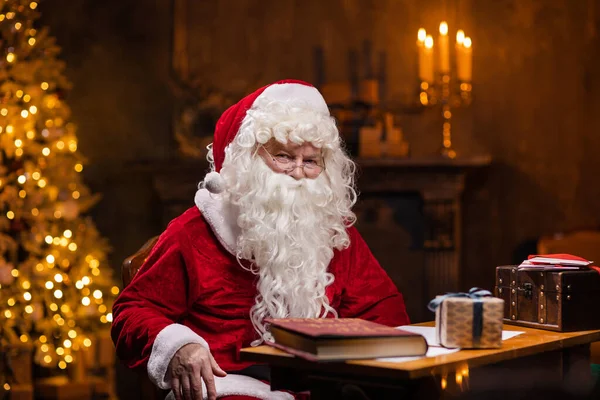サンタクロースの職場。陽気サンタはテーブルに座っている間に働いています。背景にある暖炉とクリスマスツリー。クリスマスのコンセプト. — ストック写真