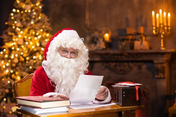 Workplace of Santa Claus. Cheerful Santa is reading letters from children while sitting at the table. Fireplace and Christmas Tree in the background. Christmas concept. Stock Picture