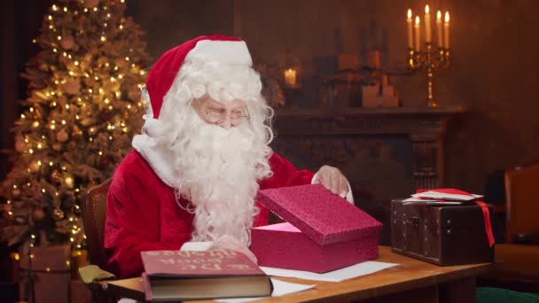 Lugar de trabajo de Santa Claus. Alegre Santa está conjurando sobre la caja de regalo mientras está sentado a la mesa. Chimenea y árbol de Navidad en el fondo. Concepto de Navidad. — Vídeos de Stock