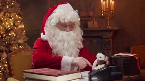 A Mikulás munkahelyén. Vidám Mikulás dolgozik a vintage telefon, miközben ül az asztalnál. Kandalló és karácsonyfa a háttérben. Karácsonyi koncepció. — Stock videók