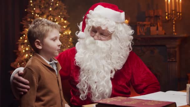 Weihnachtsmann und kleiner Junge. Der fröhliche Weihnachtsmann sitzt am Tisch und arbeitet. Kamin und Weihnachtsbaum im Hintergrund. Weihnachtskonzept. — Stockvideo