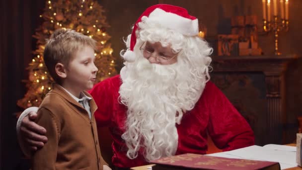 Père Noël et petit garçon. Père Noël joyeux travaille tout en étant assis à la table. Cheminée et sapin de Noël en arrière-plan. Concept de Noël. — Video