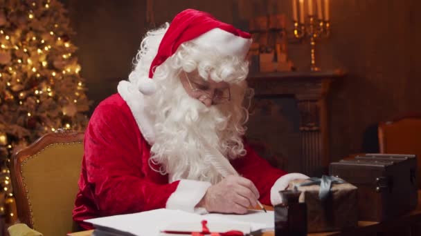 Miejsce pracy Świętego Mikołaja. Wesoły Mikołaj pisze list siedząc przy stole. Kominek i choinka w tle. Koncepcja świąteczna. — Wideo stockowe