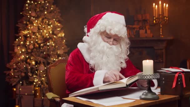 Lugar de trabajo de Santa Claus. Alegre Santa está leyendo el libro de deseos mientras está sentado a la mesa. Chimenea y árbol de Navidad en el fondo. Concepto de Navidad. — Vídeos de Stock