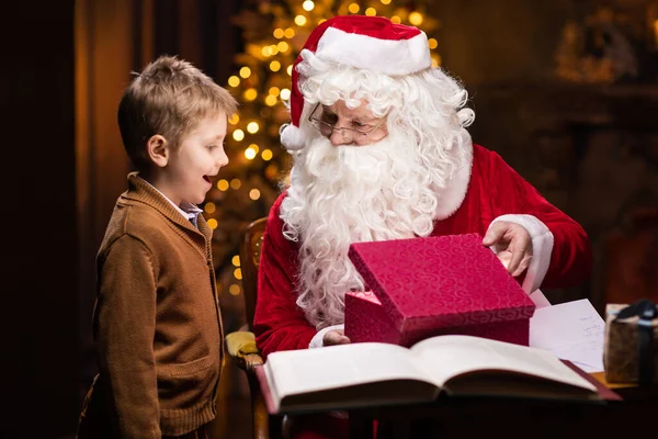 Weihnachtsmann und kleiner Junge. Der fröhliche Weihnachtsmann sitzt am Tisch und arbeitet. Kamin und Weihnachtsbaum im Hintergrund. Weihnachtskonzept. — Stockfoto