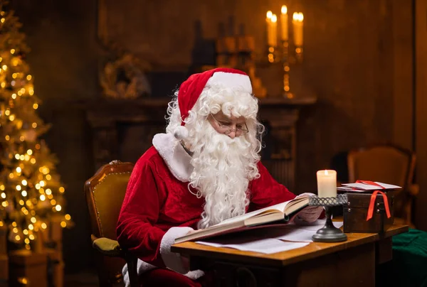 Noel Baba 'nın işyeri. Neşeli Noel Baba masada otururken dilek kitabını okuyor. Arka planda şömine ve Noel ağacı var. Noel konsepti. — Stok fotoğraf