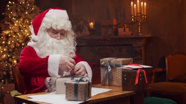 Χώρος εργασίας του Άγιου Βασίλη. Χαρούμενος Άγιος Βασίλης τυλίγει το δώρο, ενώ κάθεται στο τραπέζι. Τζάκι και χριστουγεννιάτικο δέντρο στο παρασκήνιο. Χριστουγεννιάτικη ιδέα. — Αρχείο Βίντεο