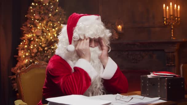 圣诞老人的工作场所。快乐的圣诞老人坐在餐桌前，一边看孩子们的来信。背景是萤火虫和圣诞树。圣诞节的概念. — 图库视频影像