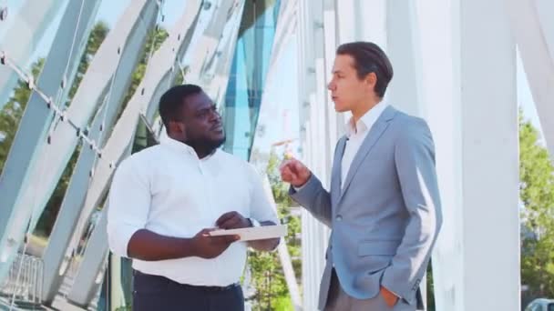 Un hombre de negocios afroamericano confiado y su colega frente al moderno edificio de oficinas. Los inversores financieros están hablando al aire libre. Banca y negocios. — Vídeos de Stock