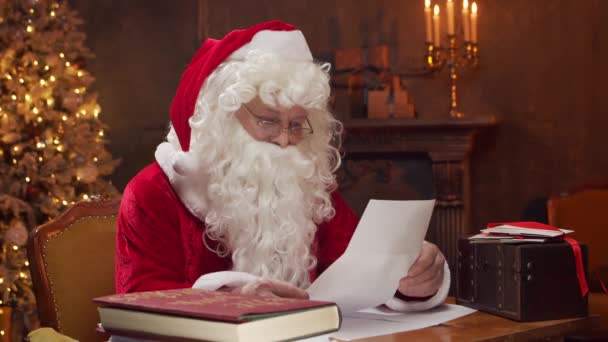 Local de trabalho do Pai Natal. O Pai Natal alegre está lendo cartas de crianças enquanto está sentado à mesa. Lareira e Árvore de Natal no fundo. Conceito de Natal. — Vídeo de Stock