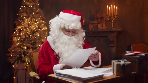 サンタクロースの職場。陽気サンタはテーブルに座っている間に子供たちからの手紙を読んでいます。背景にある暖炉とクリスマスツリー。クリスマスのコンセプト. — ストック動画