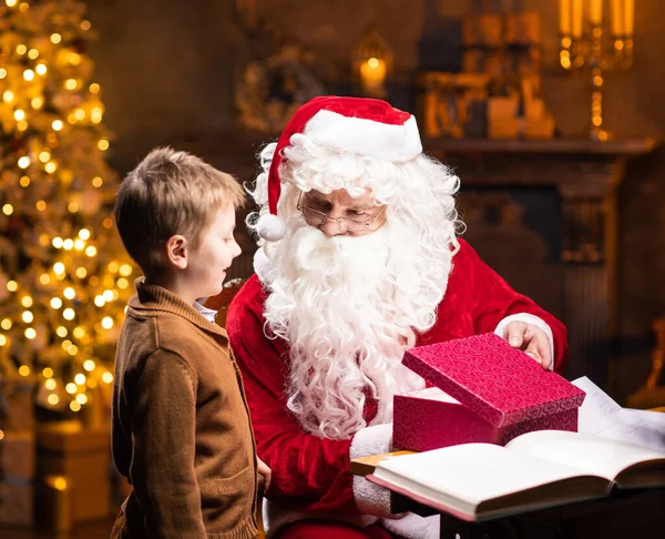 Weihnachtsmann und kleiner Junge. Der fröhliche Weihnachtsmann sitzt am Tisch und arbeitet. Kamin und Weihnachtsbaum im Hintergrund. Weihnachtskonzept. — Stockfoto