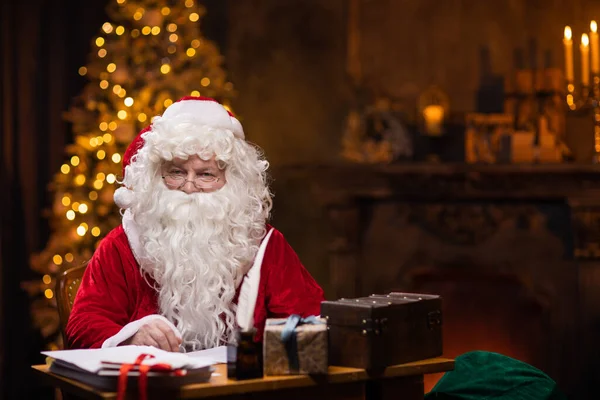 Workplace of Santa Claus. Cheerful Santa is working while sitting at the table. Fireplace and Christmas Tree in the background. Christmas concept. — Stock Photo, Image