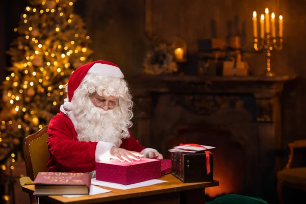 サンタクロースの職場。陽気サンタはテーブルに座っている間にギフトボックスの上に集まっています。背景にある暖炉とクリスマスツリー。クリスマスのコンセプト. — ストック写真