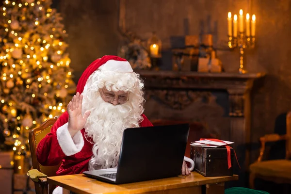 Lugar de trabajo de Santa Claus. Alegre Santa está trabajando en el portátil mientras está sentado en la mesa. Chimenea y árbol de Navidad en el fondo. Concepto de Navidad. —  Fotos de Stock