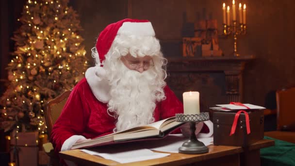 A Mikulás munkahelyén. Vidám Mikulás olvassa a kívánságok könyvét, miközben ül az asztalnál. Kandalló és karácsonyfa a háttérben. Karácsonyi koncepció. — Stock videók