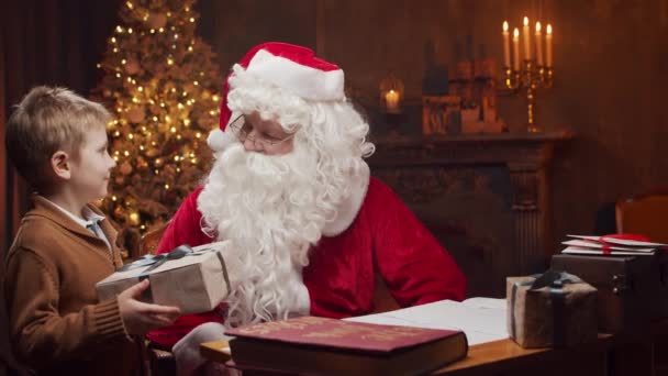 Santa Claus y el niño pequeño. Alegre Santa está trabajando mientras está sentado a la mesa. Chimenea y árbol de Navidad en el fondo. Concepto de Navidad. — Vídeos de Stock