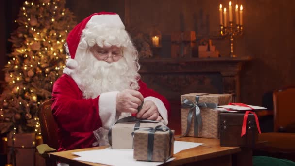 Lugar de trabajo de Santa Claus. Alegre Santa está envolviendo el regalo mientras está sentado en la mesa. Chimenea y árbol de Navidad en el fondo. Concepto de Navidad. — Vídeos de Stock