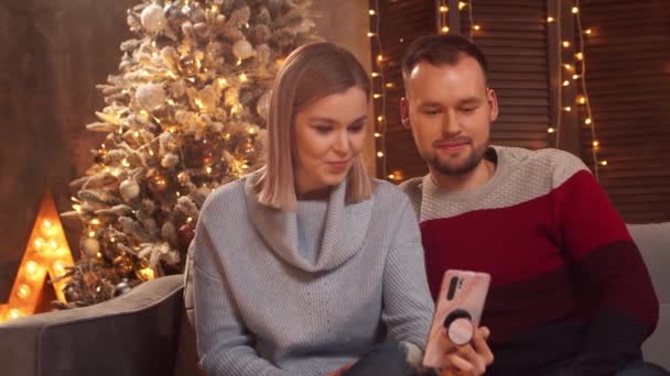 Jeune couple amoureux célébrant Noël à la maison. Jeune homme et sa petite amie dans un intérieur de maison avec un arbre de Noël sur le fond. — Video