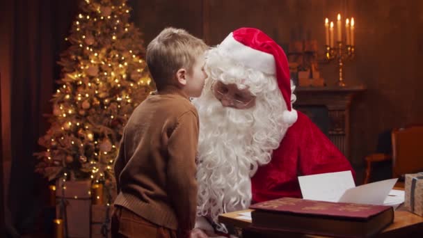 Santa Claus y el niño pequeño. Alegre Santa está trabajando mientras está sentado a la mesa. Chimenea y árbol de Navidad en el fondo. Concepto de Navidad. — Vídeos de Stock