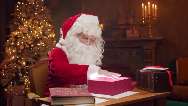 Lugar de trabajo de Santa Claus. Alegre Santa está conjurando sobre la caja de regalo mientras está sentado a la mesa. Chimenea y árbol de Navidad en el fondo. Concepto de Navidad. — Vídeos de Stock