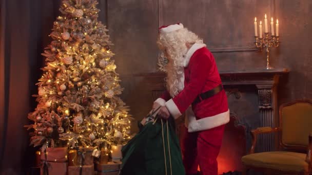 Papai Noel coloca presentes sob a árvore de Natal de sua bolsa. Interior da casa com lareira e velas. Conceito de Natal. — Vídeo de Stock