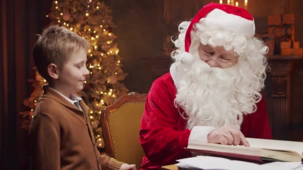 Pai Natal e rapazinho. O Pai Natal alegre está a trabalhar enquanto está sentado à mesa. Lareira e Árvore de Natal no fundo. Conceito de Natal. — Vídeo de Stock