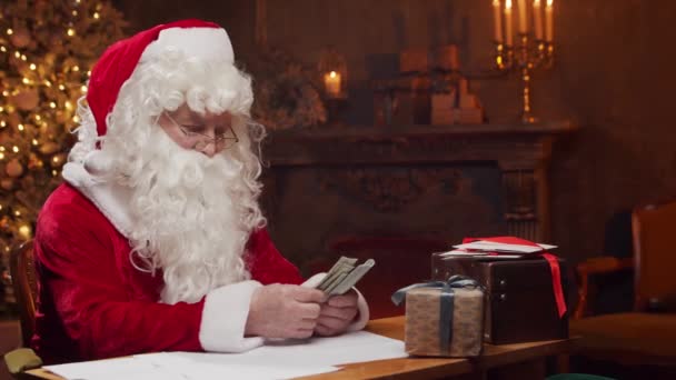 Lieu de travail du Père Noël. Père Noël joyeux compte de l'argent tout en étant assis à la table. Cheminée et sapin de Noël en arrière-plan. Concept de Noël. — Video