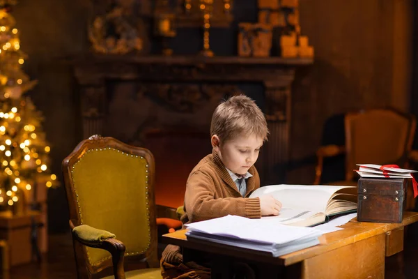 Pojken läser en magisk bok när han sitter vid bordet. Heminredning med julgran och öppen spis. Traditionellt julkoncept. — Stockfoto