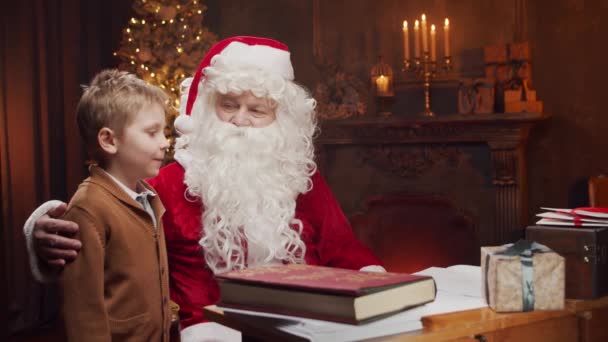 Santa Claus y el niño pequeño. Alegre Santa está trabajando mientras está sentado a la mesa. Chimenea y árbol de Navidad en el fondo. Concepto de Navidad. — Vídeos de Stock