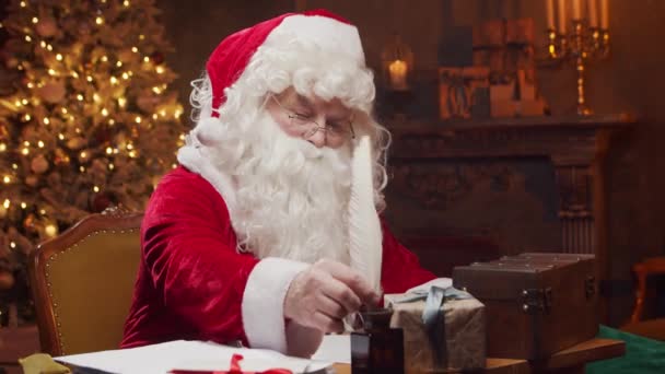 Lugar de trabajo de Santa Claus. Alegre Santa está escribiendo la carta mientras está sentado a la mesa. Chimenea y árbol de Navidad en el fondo. Concepto de Navidad. — Vídeos de Stock
