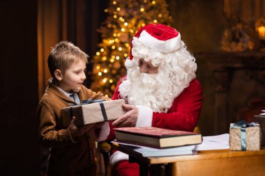 Noel Baba ve küçük çocuk. Neşeli Noel Baba masada otururken çalışıyor. Arka planda şömine ve Noel ağacı var. Noel konsepti.