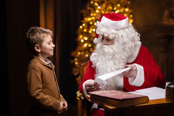 Père Noël et petit garçon. Père Noël joyeux travaille tout en étant assis à la table. Cheminée et sapin de Noël en arrière-plan. Concept de Noël. — Photo