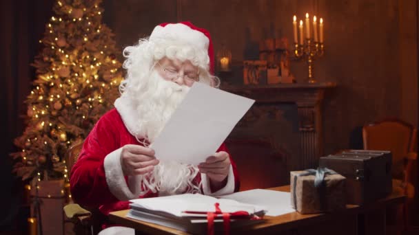 A Mikulás munkahelyén. Vidám Mikulás olvas leveleket a gyerekek, miközben ül az asztalnál. Kandalló és karácsonyfa a háttérben. Karácsonyi koncepció. — Stock videók
