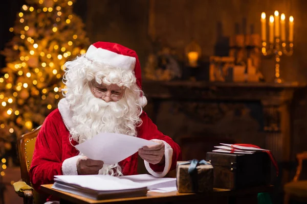 Arbeitsplatz des Weihnachtsmannes. Der fröhliche Weihnachtsmann liest am Tisch Briefe von Kindern vor. Kamin und Weihnachtsbaum im Hintergrund. Weihnachtskonzept. — Stockfoto